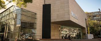 Modern building with "MALBA" on the front, glass and stone facade, trees surrounding, sleek design.