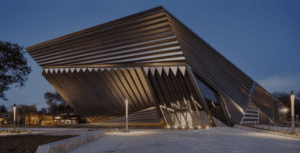 Large modern building with sharp angles and metallic exterior illuminated at dusk, surrounded by trees.