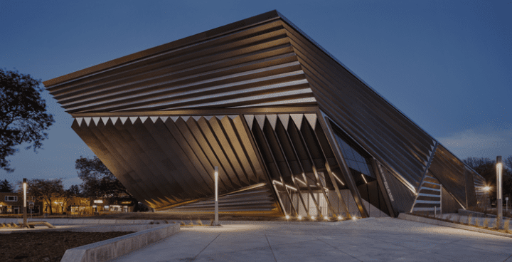 Large modern building with sharp angles and metallic exterior illuminated at dusk, surrounded by trees.