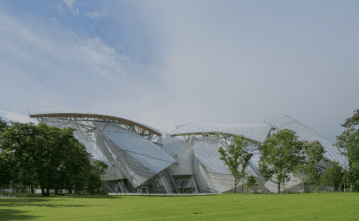 Modern metal and glass structure in a park with trees and lawn under a partly cloudy sky.