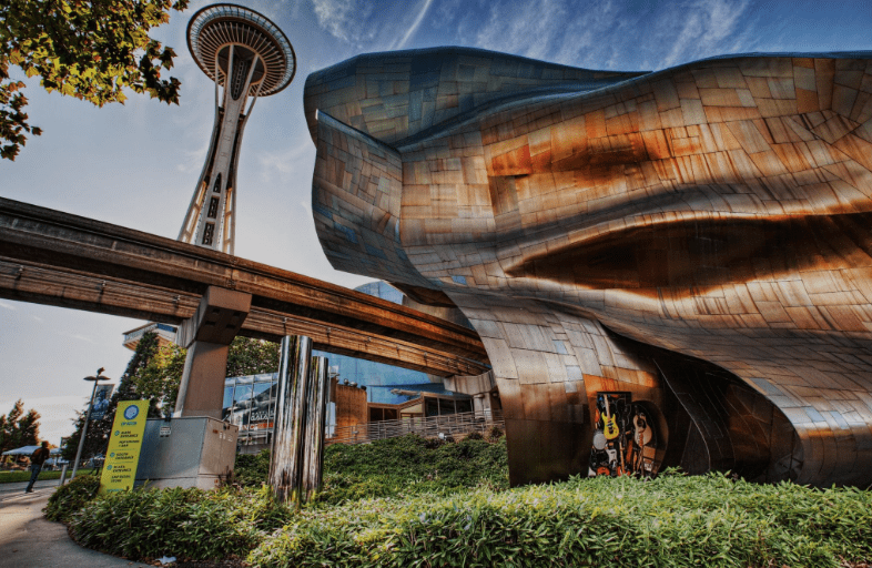 Modern wavy metallic building near the Space Needle, Seattle, with monorail and greenery in foreground