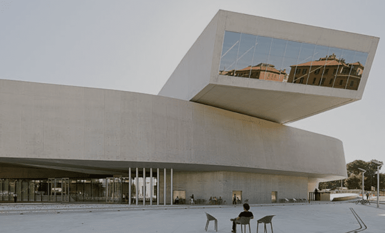 A minimalist modern building with glass windows and an overhanging structure, a person seated outside.