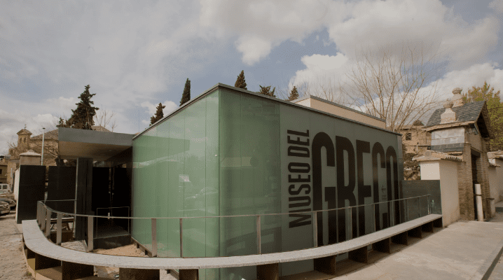 Modern building with glass walls and "Museo del Greco" sign surrounded by trees and other structures.