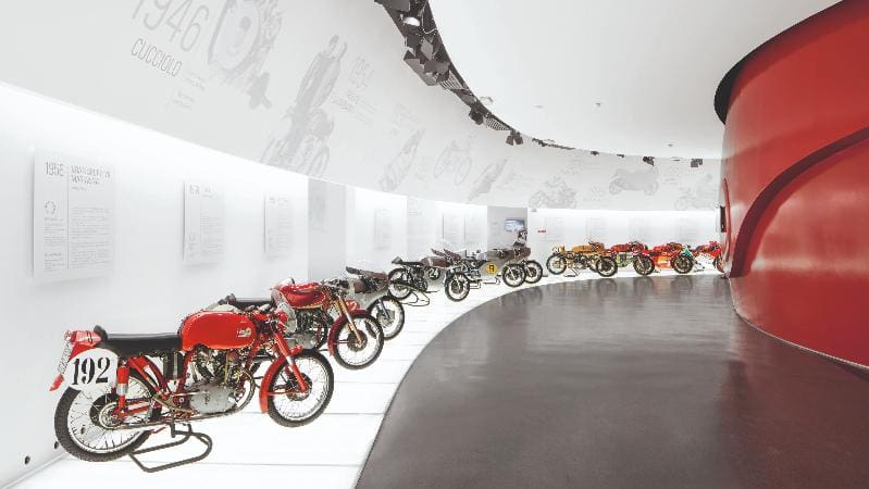Modern museum gallery with classic motorcycles displayed in a curved, white-walled corridor.