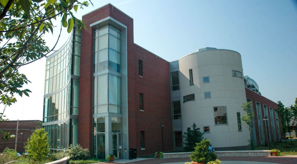 Modern red brick and light concrete building with curved glass facade, windows, and surrounding greenery.