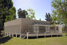 Modern single-story building with flat roof, large porch, steps, green lawn, and surrounding trees.