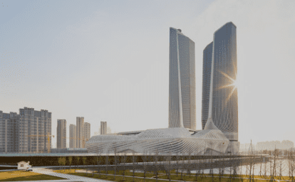 Modern architectural complex with two tall skyscrapers, sun reflecting off one, landscaped area.