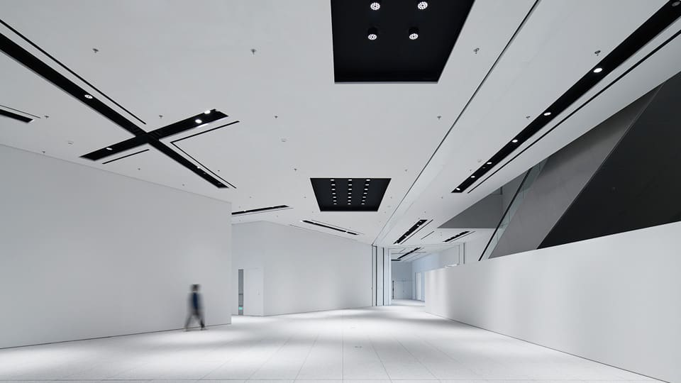 Modern spacious room with high ceilings, geometric lighting, bright walls, and blurred figure walking.