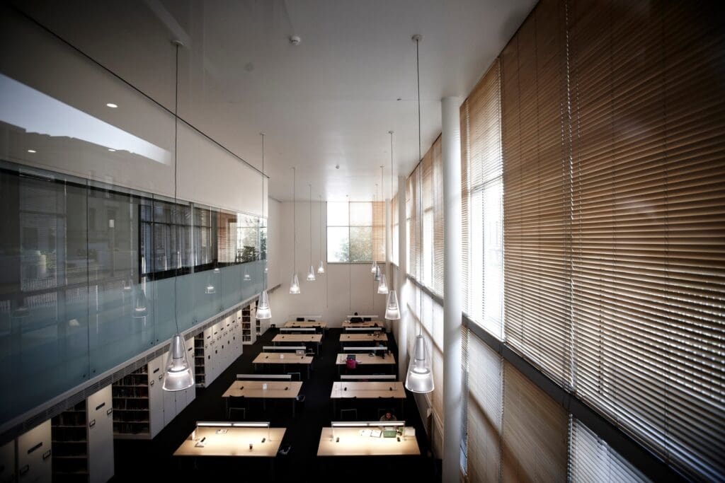 Modern, spacious room with high ceiling, pendant lights, desks, large windows with blinds, and bookshelves.