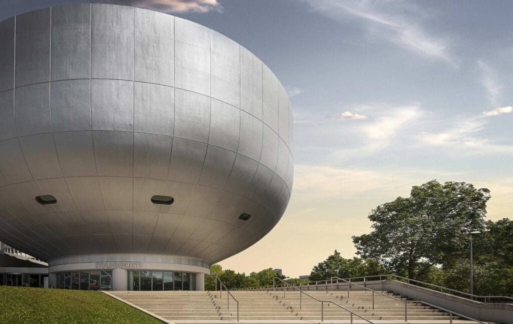Modern spherical museum with metallic exterior, stairs, and surrounding greenery under a partly cloudy sky.