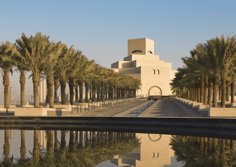 Modern beige stone building with palm-lined pathway and reflective pool under clear sky, urban skyline.