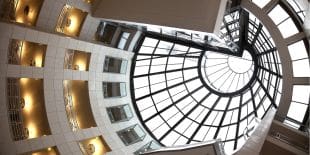 Upward view through a modern structure with a circular glass dome and tiered floors with lit alcoves