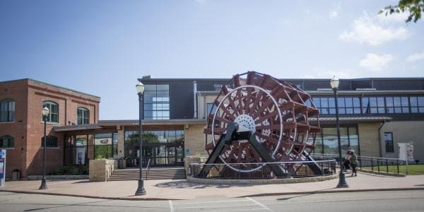 Building with large windows, a waterwheel, stone pillars, and lampposts showing modern and traditional architecture.