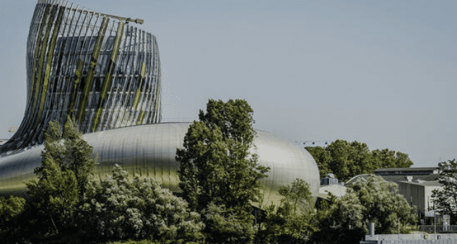 Modern cylindrical building with twisted design, metallic surfaces, and windows surrounded by trees.