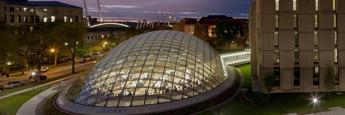 Modern urban library with dome-shaped glass roof, illuminated at dusk, surrounded by buildings and trees