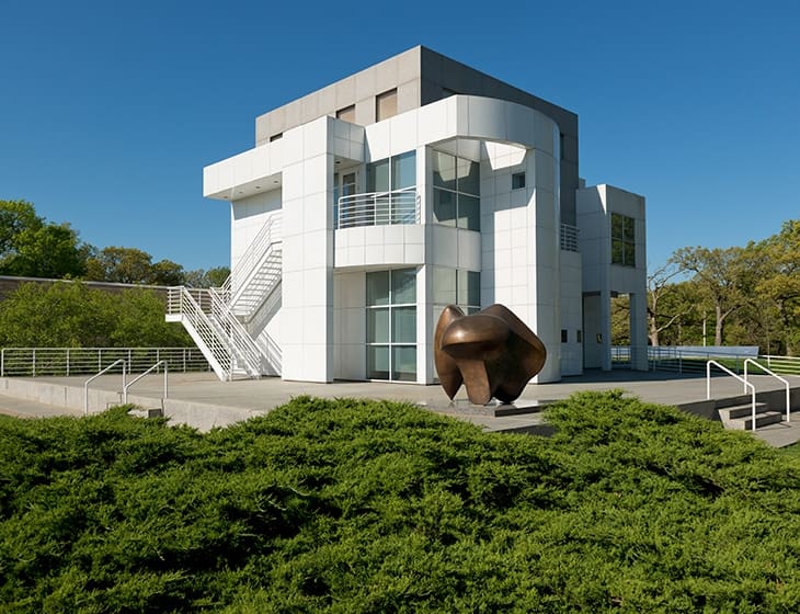 Modern two-story white building with large glass windows, balconies, and outdoor staircase, surrounded by greenery.