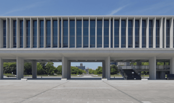 Modernist building with vertical windows, open ground-level area, pathway, and greenery under clear sky