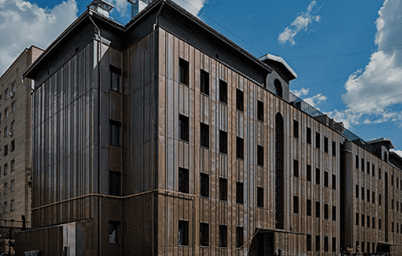 Multi-story building with brown panel exterior, many windows, multifaceted roof, and chimneys.