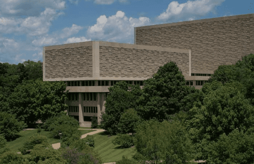 Multi-story rectangular building partly obscured by green foliage, with pathways and a partly cloudy sky.