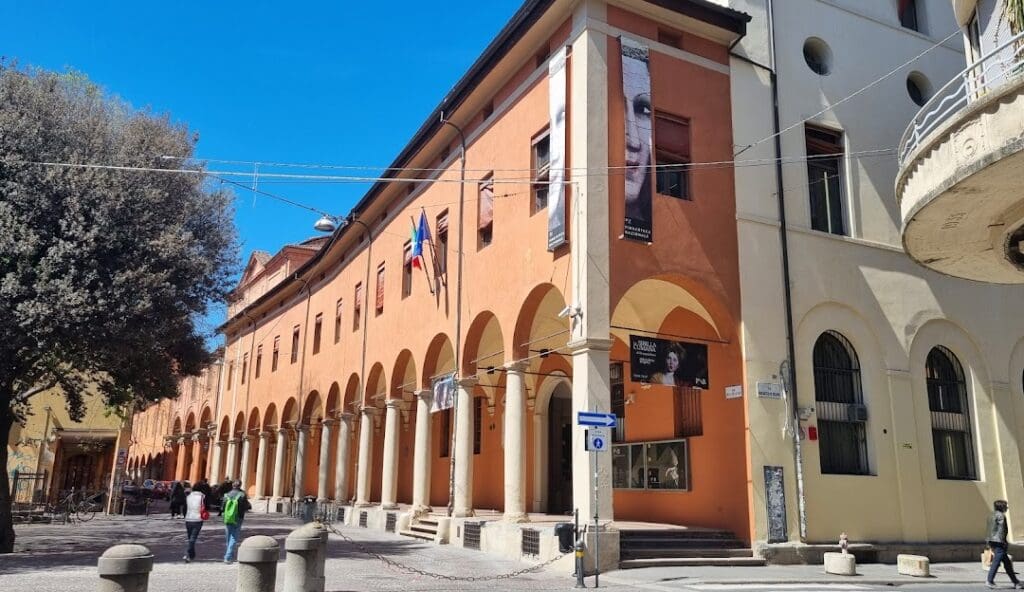 Multi-story orange building with arched columns, banners, flags, and pedestrians under blue sky.