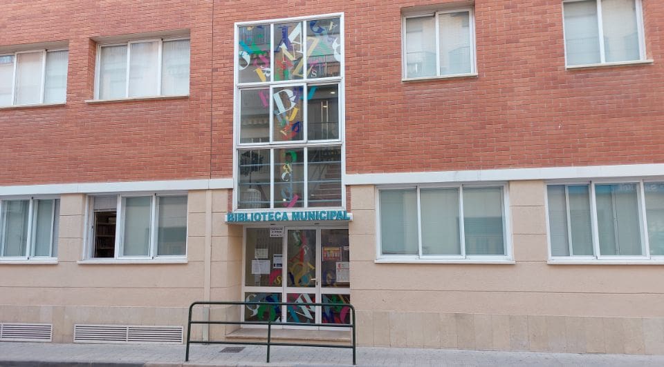 Municipal library exterior with red brick facade, glass entrance with colorful designs, and a sign.