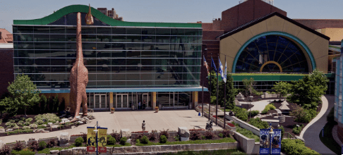 Museum entrance with tall dinosaur sculpture, modern glass façade, dome structure, and landscaped gardens