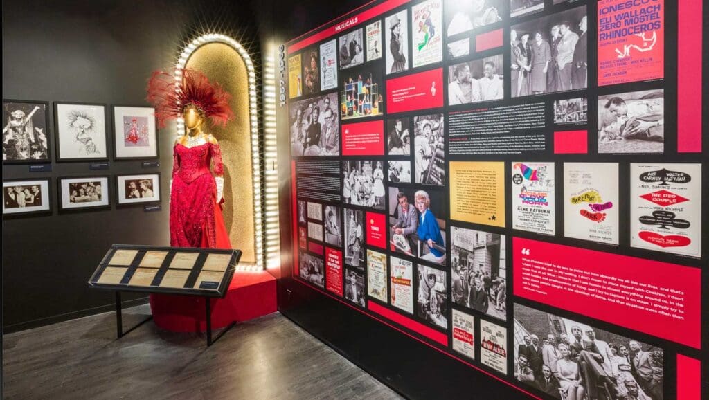 Museum exhibit with a red costume, feathers, beads, photos, posters, texts, and a document table about musicals.