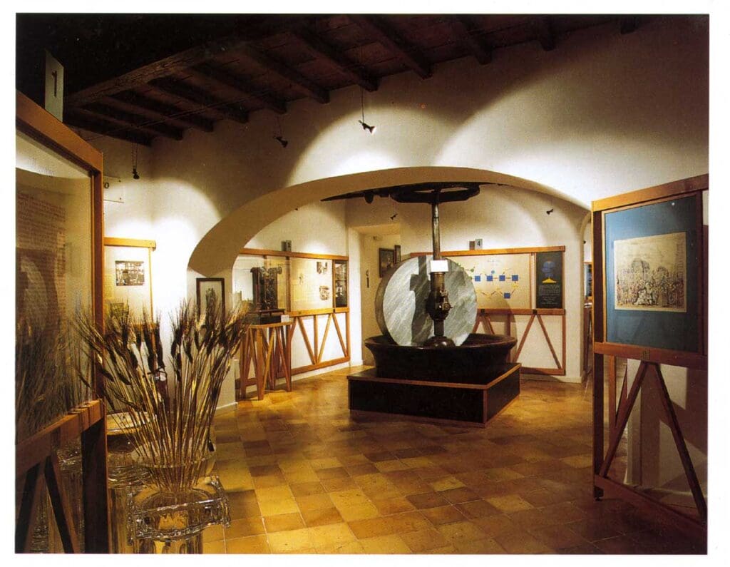 Museum exhibition room with framed artworks, historical items, a circular mechanical device, and wheat bundles.