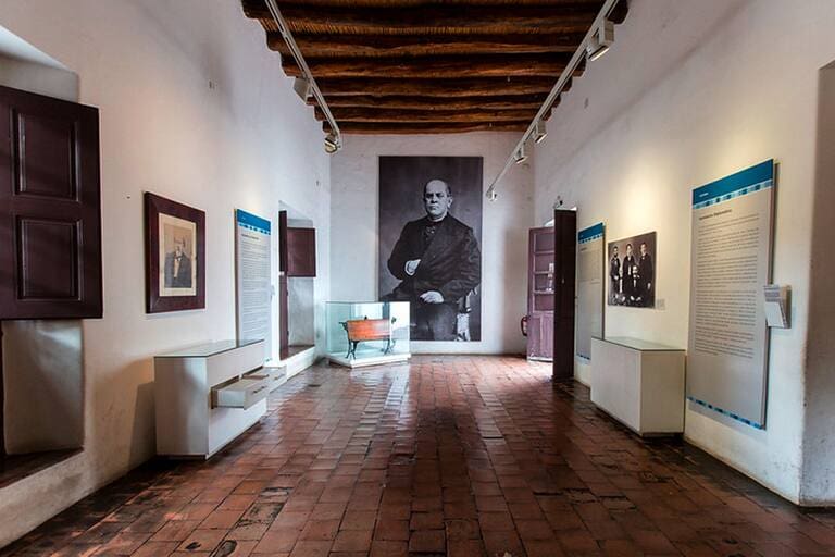 Museum gallery with white walls, brick floor, large historical portrait, display cases, and wooden beam ceiling