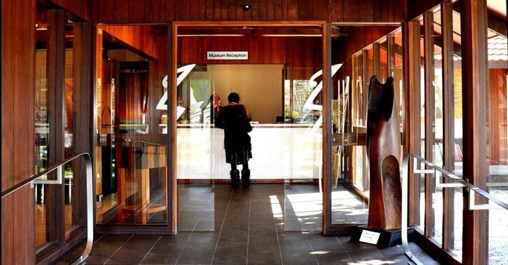 Entrance to museum reception with wooden walls, glass doors, a person at desk, and a wooden sculpture.