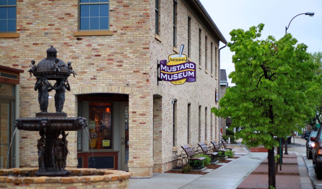 Two-story brick building labeled National Mustard Museum, with a fountain, trees, benches, and sidewalk.