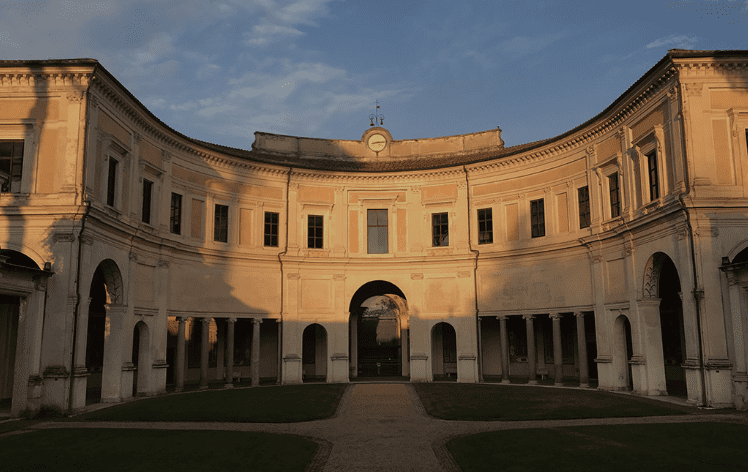 Curved neoclassical building with arched doorways, clock, clear sky, grassy courtyard, central pathway.