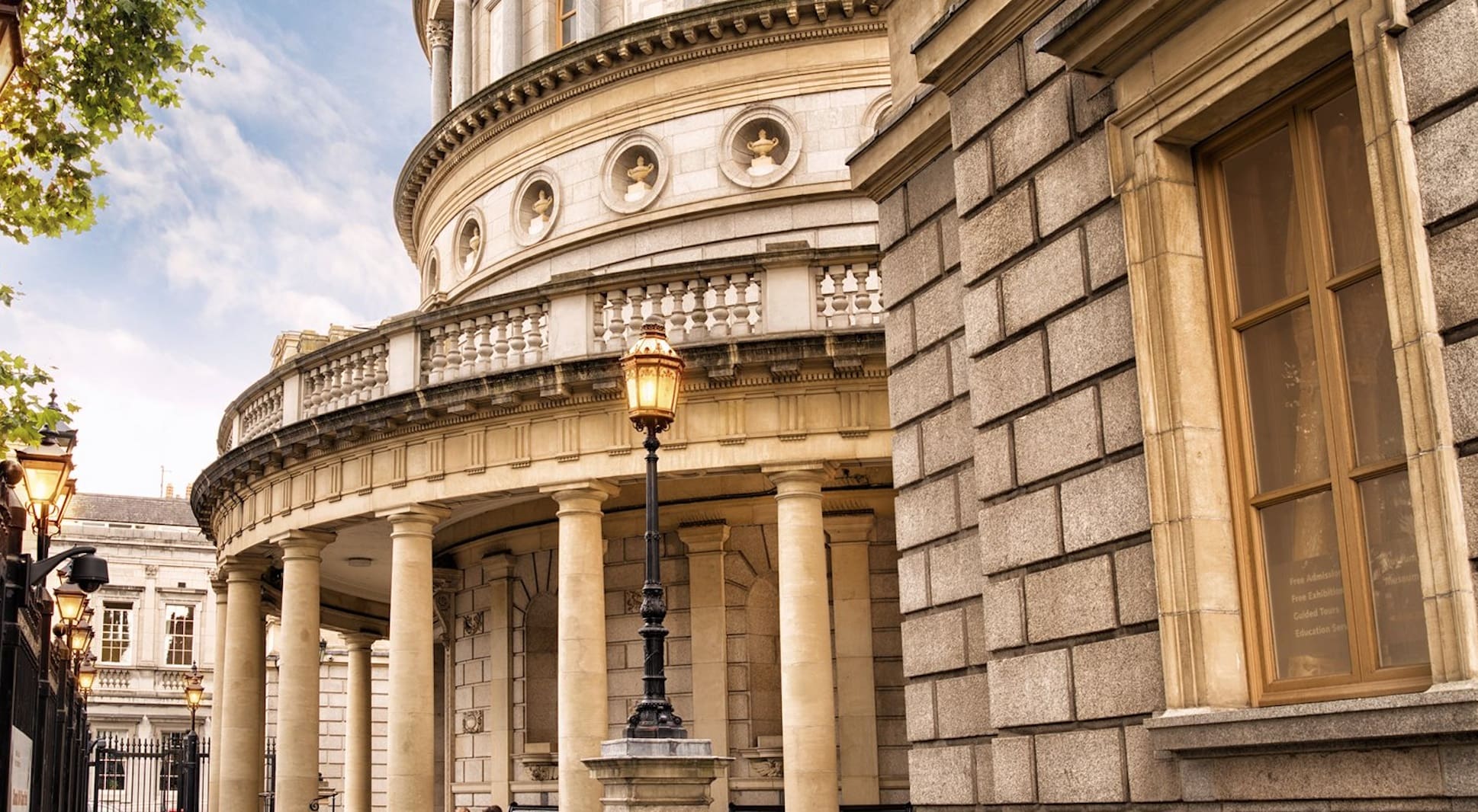 Neoclassical building with columns, ornate reliefs, and a lamppost under a partly cloudy sky.