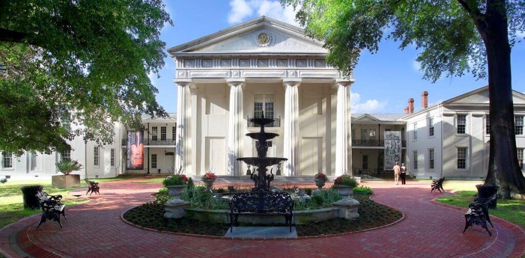 Neoclassical building with tall columns, a courtyard fountain, benches, trees, and clear blue sky.