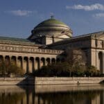 Neoclassical building with central dome and portico, reflected in water, surrounded by trees.