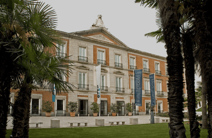 Large neoclassical building with banners, palm trees, outdoor seating, and manicured lawn in front.