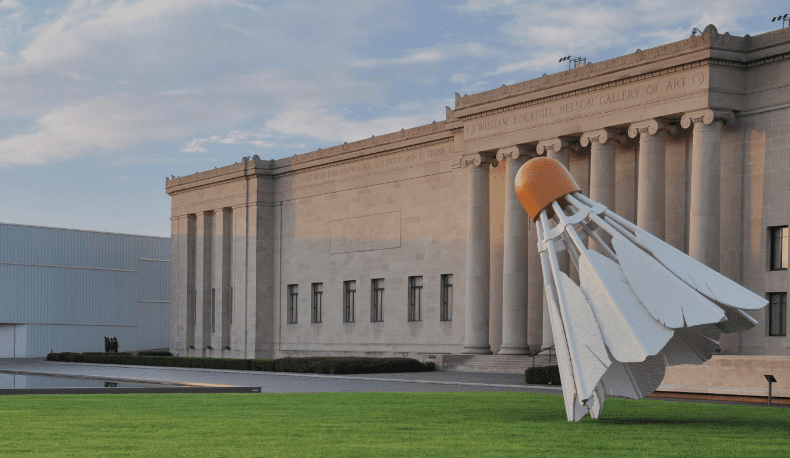 Neoclassical building with columns and a large shuttlecock sculpture on a grassy lawn under partly cloudy sky.
