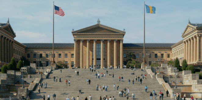 Neoclassical building with staircase, columns, two flags, and people on steps in sunny weather