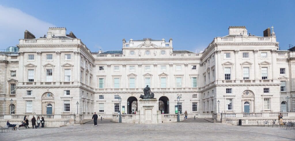A large neoclassical building with columns, a central statue, and a courtyard with people and lampposts.