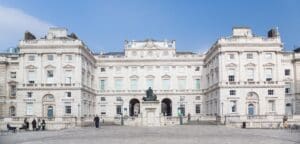 A large neoclassical building with columns, a central statue, and a courtyard with people and lampposts.