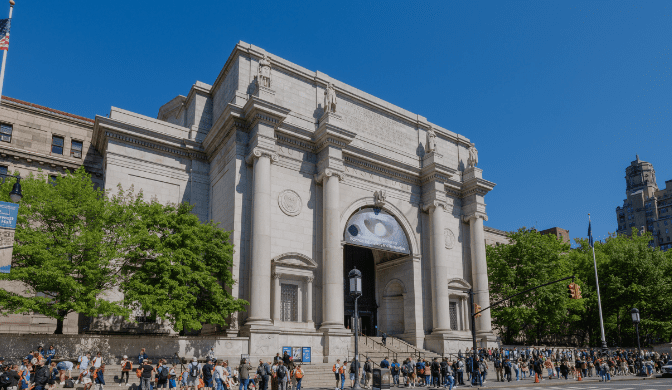 Neoclassical building with tall columns, arches, people gathered outside, trees, and clear blue sky.