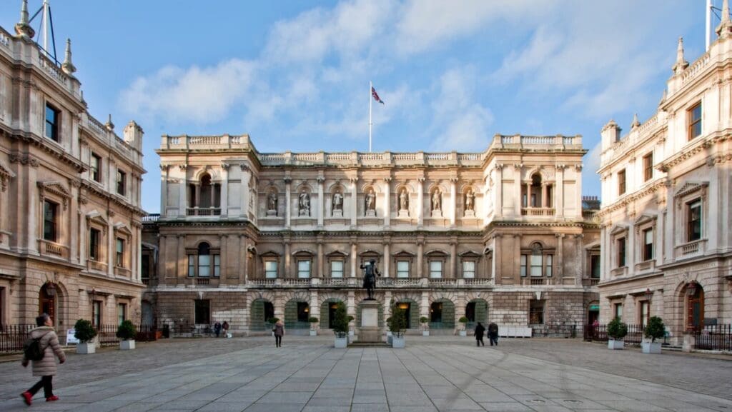 Neoclassical courtyard with central statue, symmetrical building, alcove statues, flag, people, and plants.