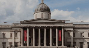 Neoclassical gallery with a large dome, columned facade, red banners, and partly cloudy sky.