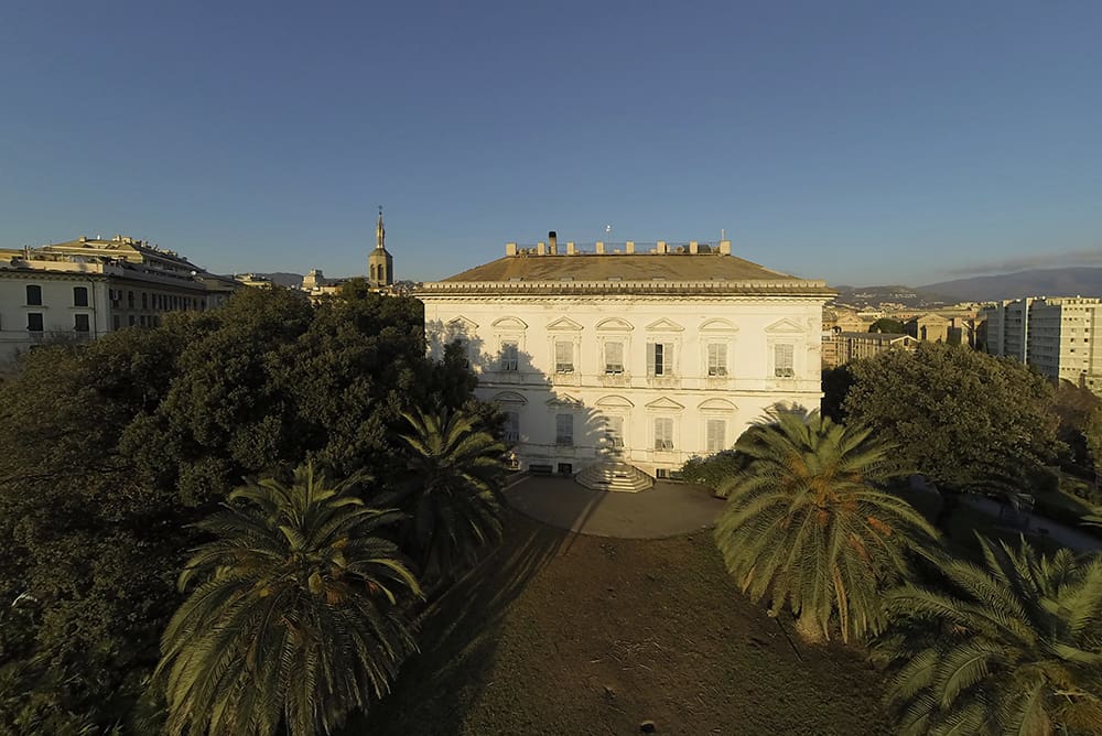 Neoclassical white building with symmetrical windows, surrounded by trees, palms, and nearby cityscape.