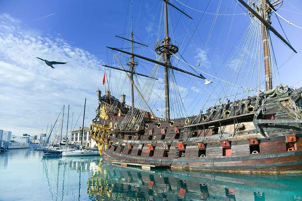 Old-style wooden sailing ship docked in a calm harbor, surrounded by modern boats and flying seagulls