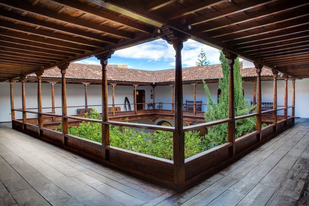 Open courtyard with wooden veranda, railings, columns, tiled roof, white walls, and lush garden under clear sky