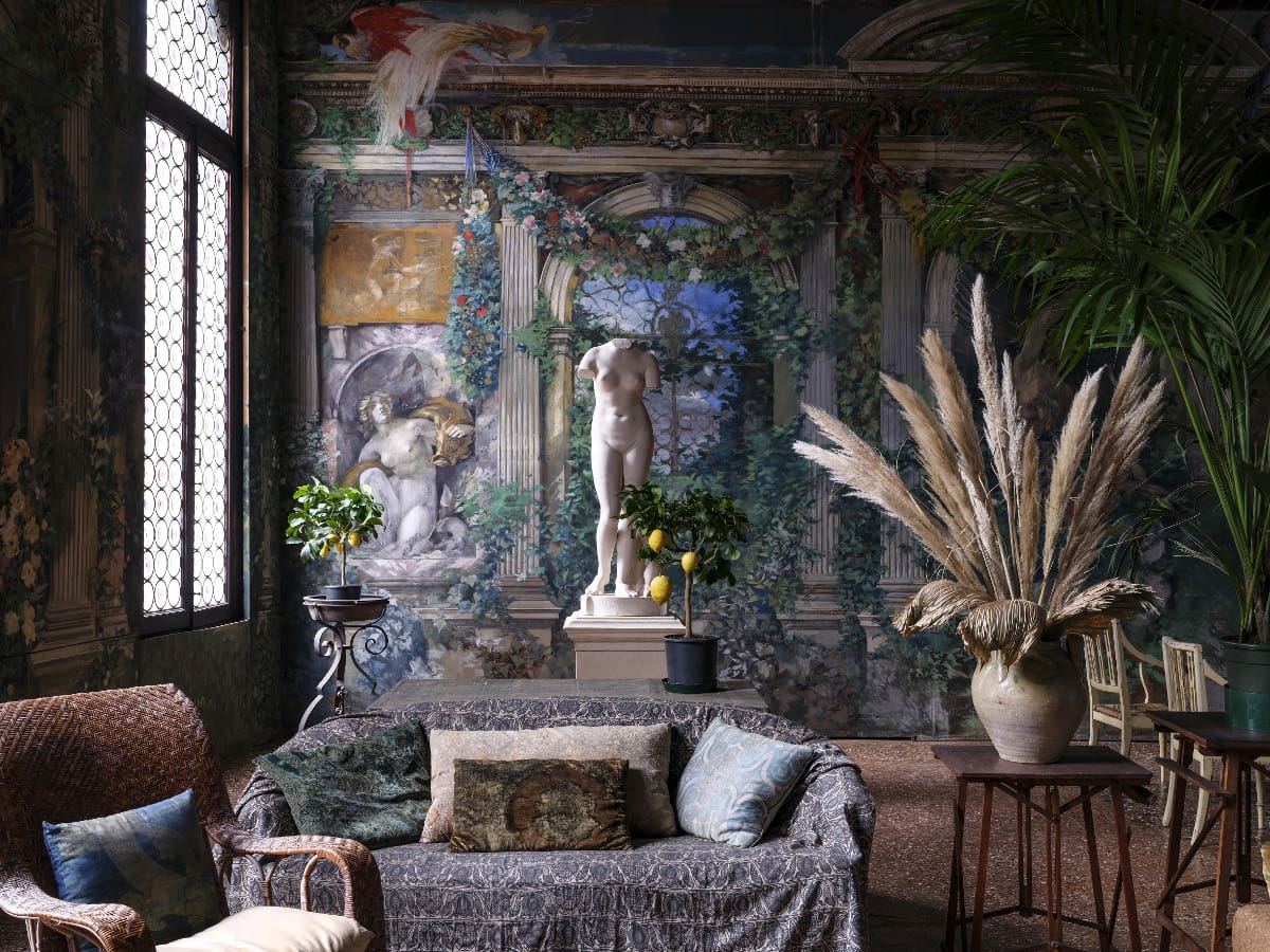 Opulent indoor space with classical murals, a central statue, plants, and rustic furniture.