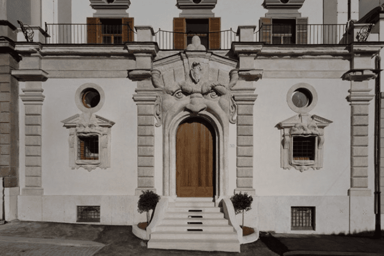 Building facade with ornate entrance featuring a large face sculpture, wooden door, and flanking trees