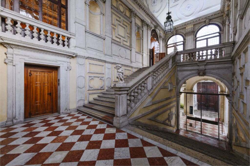 Grand interior with ornate staircase, intricate railings, statue, and decorative moldings by arched windows.