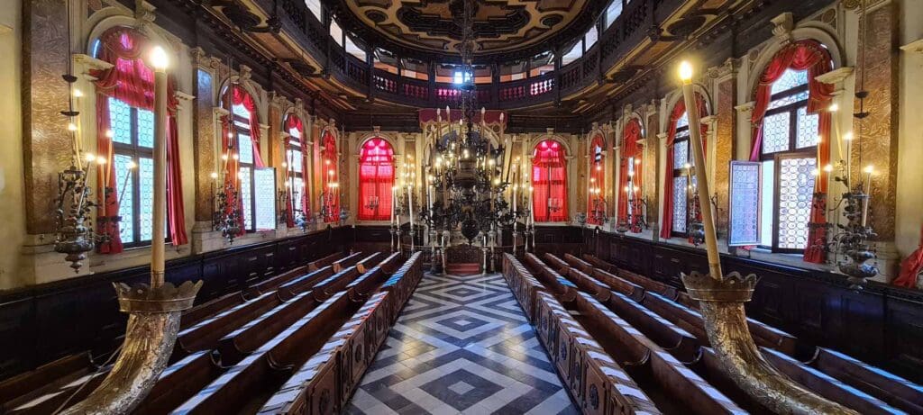 Ornate historic building interior with chandeliers, red-draped windows, wooden pews, and a grand altar.